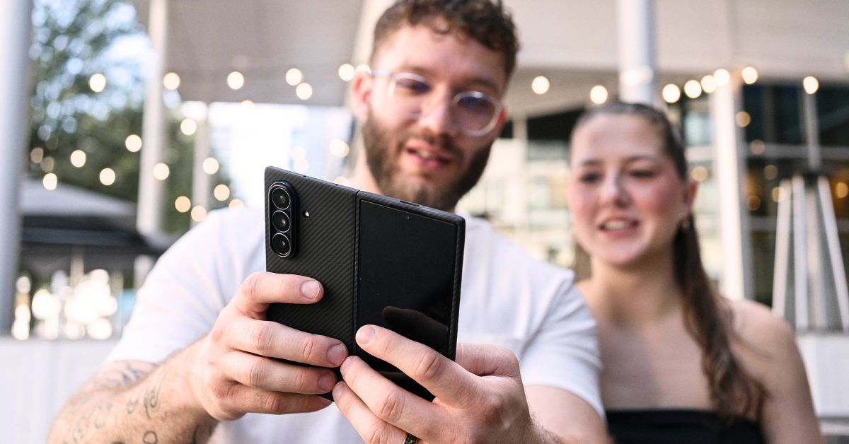 黒いカーボン調のGalaxy Z Fold 7ケースを持ち、自撮りをする人物(背景にも人物が写っている)