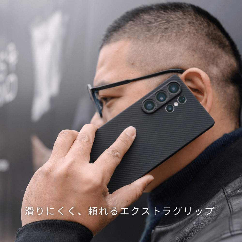  黒いカーボン調のSamsung Galaxy S25 Ultra ケースを装着したスマホで電話をしている男性。下に「滑りにくく、頼れるエクストラグリップ」と書かれています。
